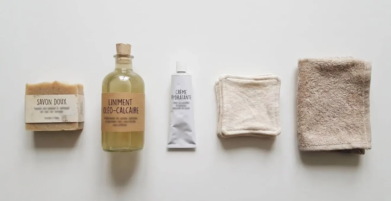 Vue en plongée sur une sélection minimaliste de 5 produits de soins pour bébé alignés sur fond blanc : savon, liniment, crème, coton lavable et serviette